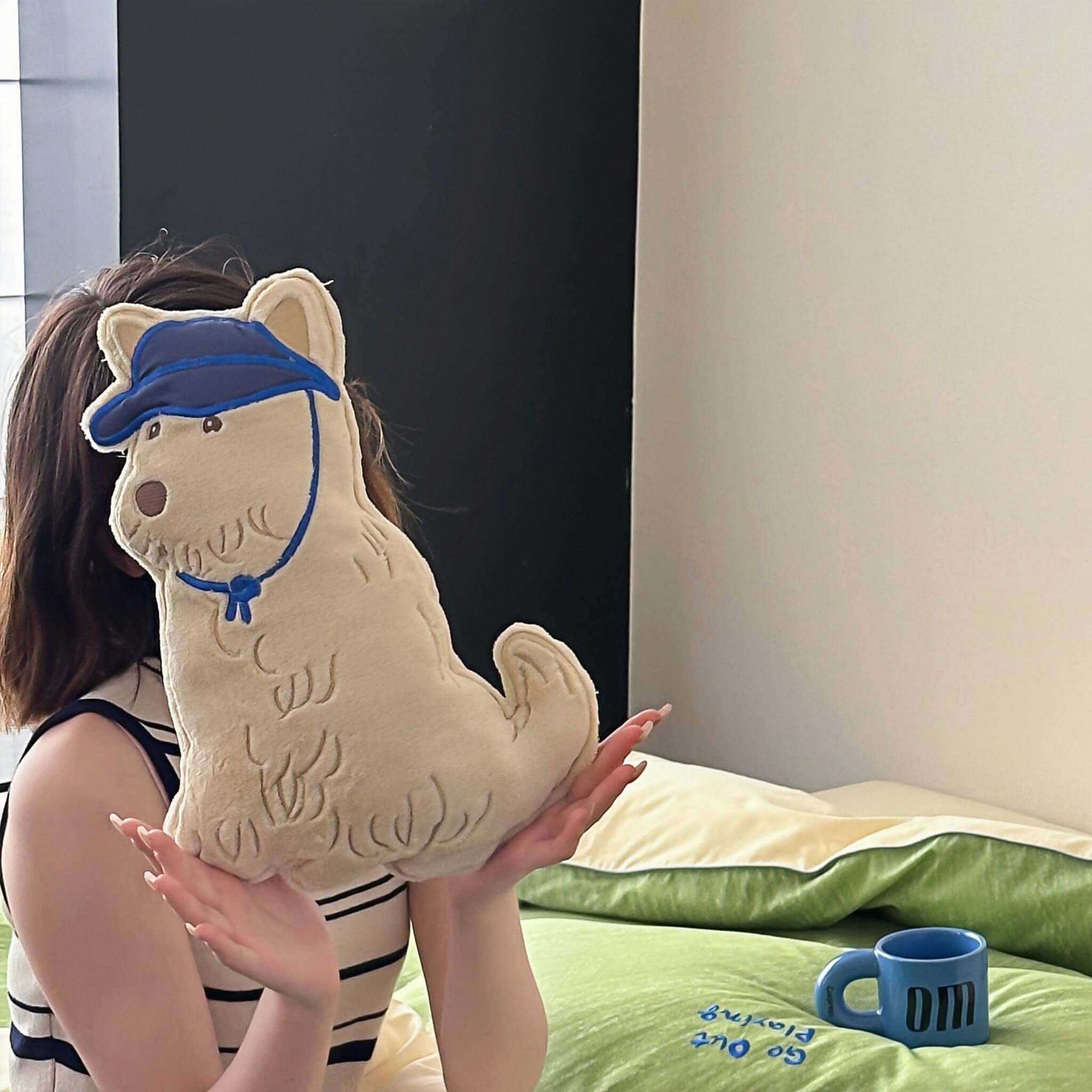 Person holding a plush dog toy with a blue cap in a room with green bedding.