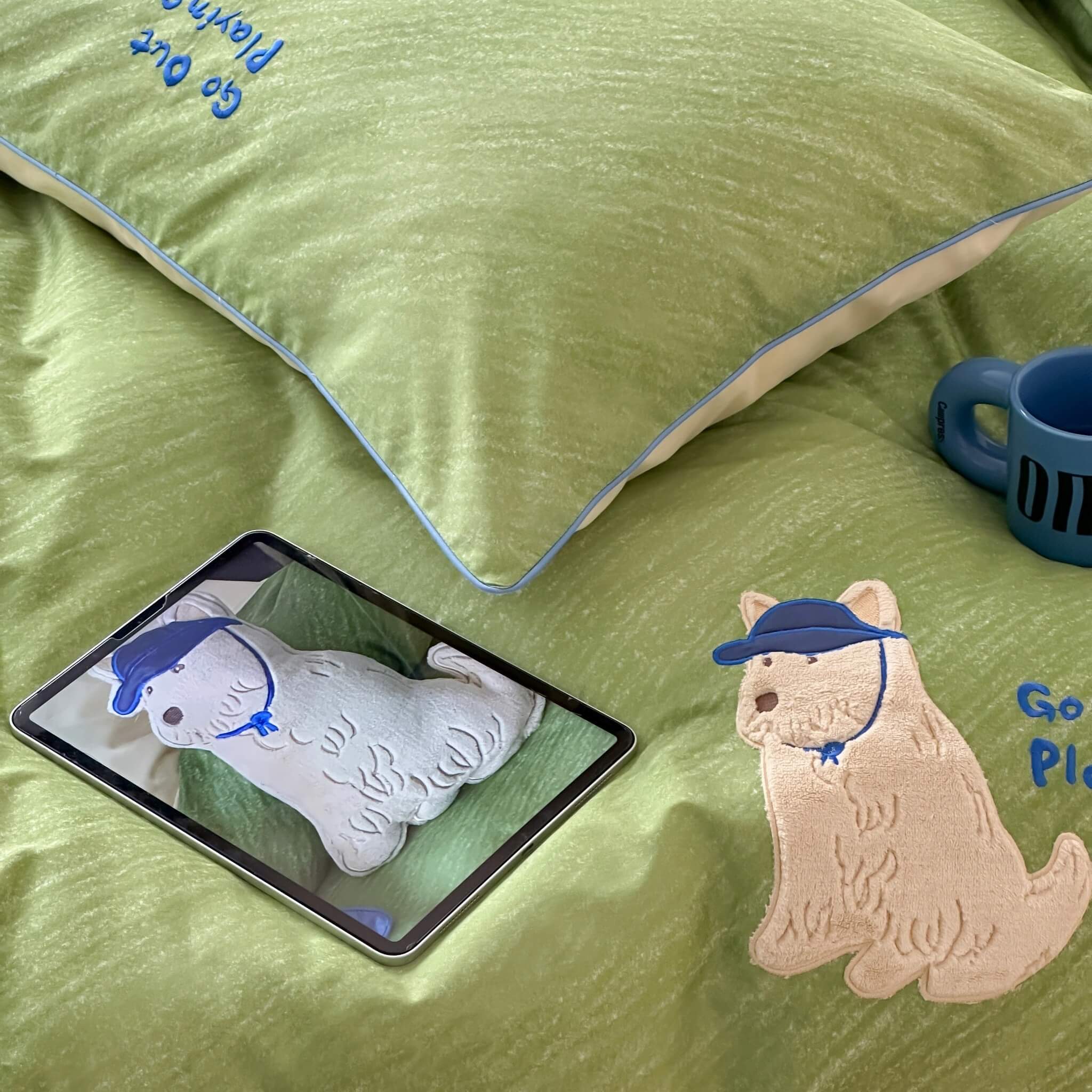 Green bedspread with embroidered dog design, tablet displaying similar design, and blue mug.