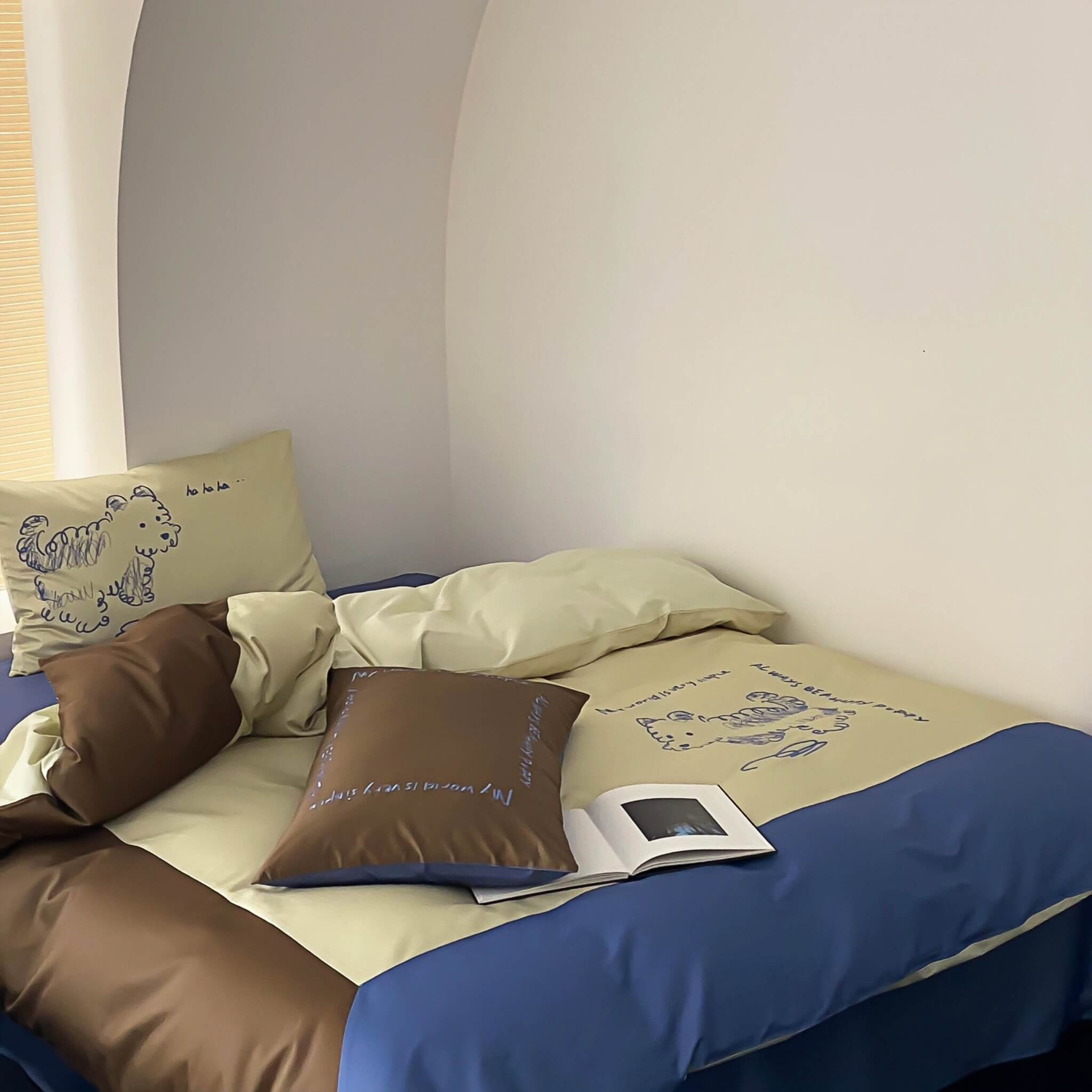 Bed with blue and beige bedding, brown pillows, embroided dog design,  and an open book.