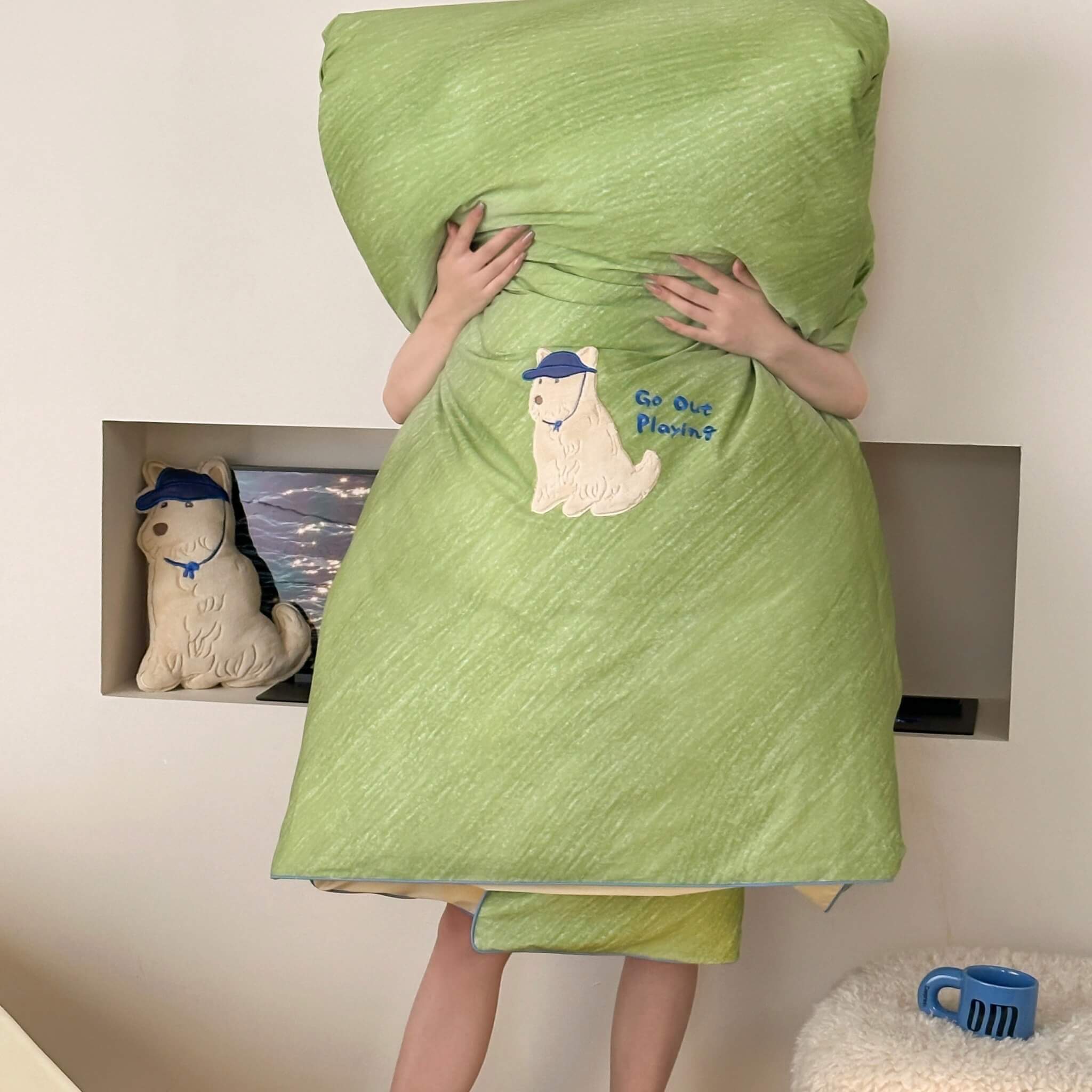 Person wrapped in a green quilt with a cartoon dog character, standing in front of a wall with decorative items.