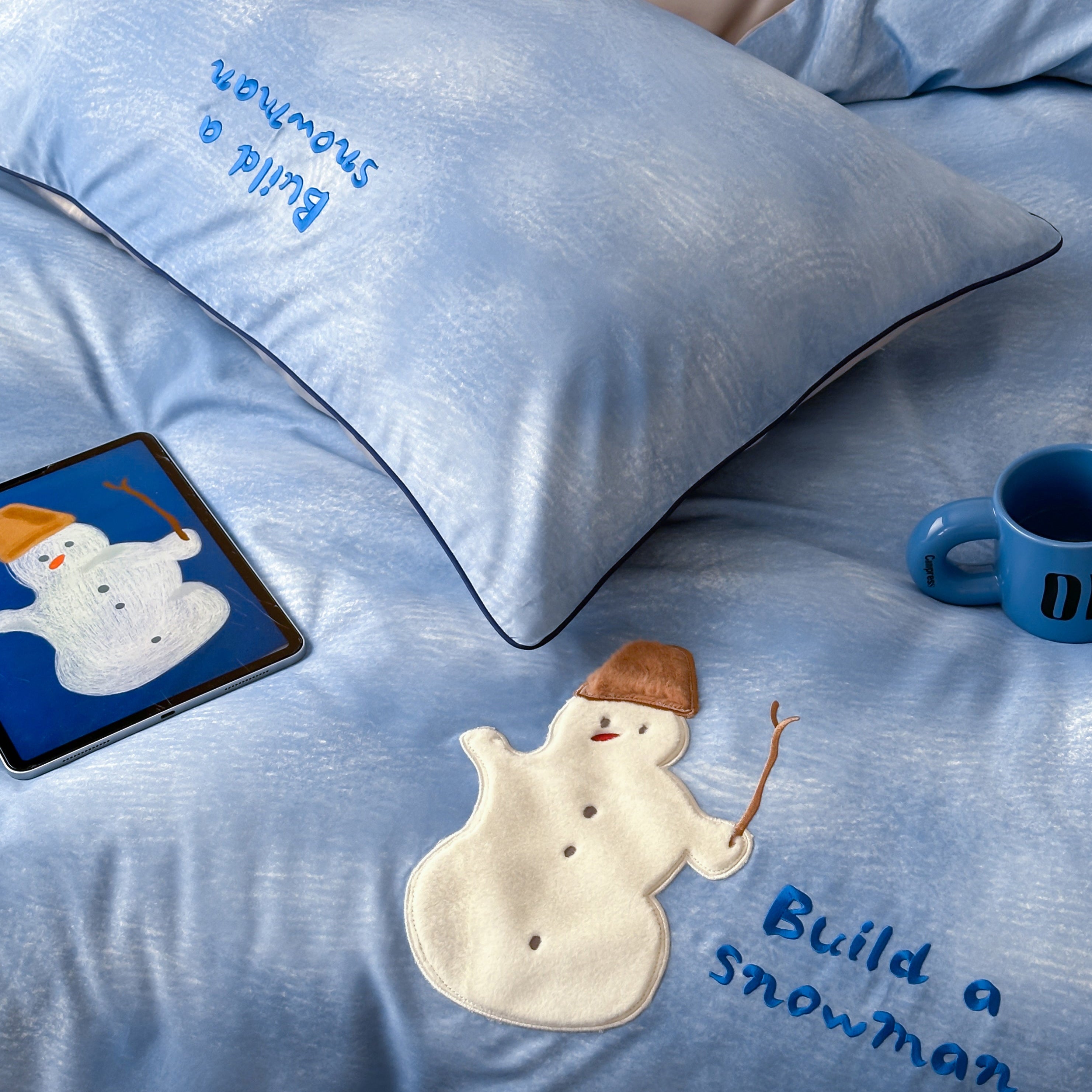 Blue bedding set with snowman design and 'Build a Snowman' text, tablet, and mug on a bed.